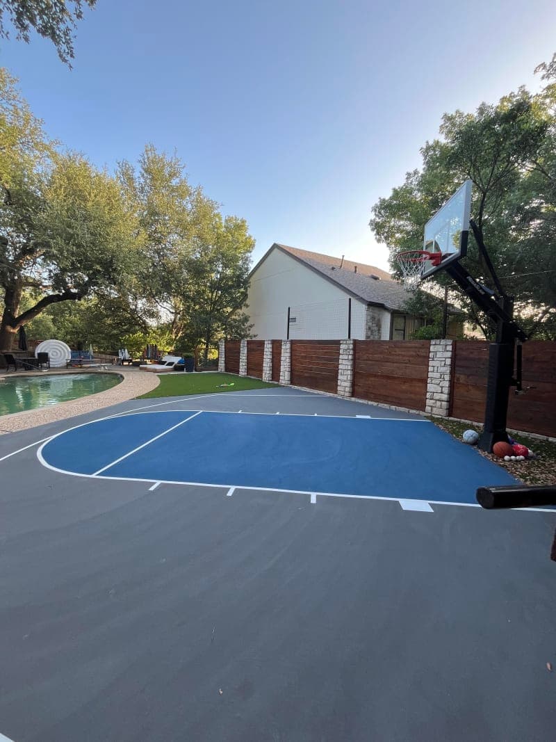Finished blue and gray residential backyard basketball court next to a pool with a stone and wood fence in Austin, Texas